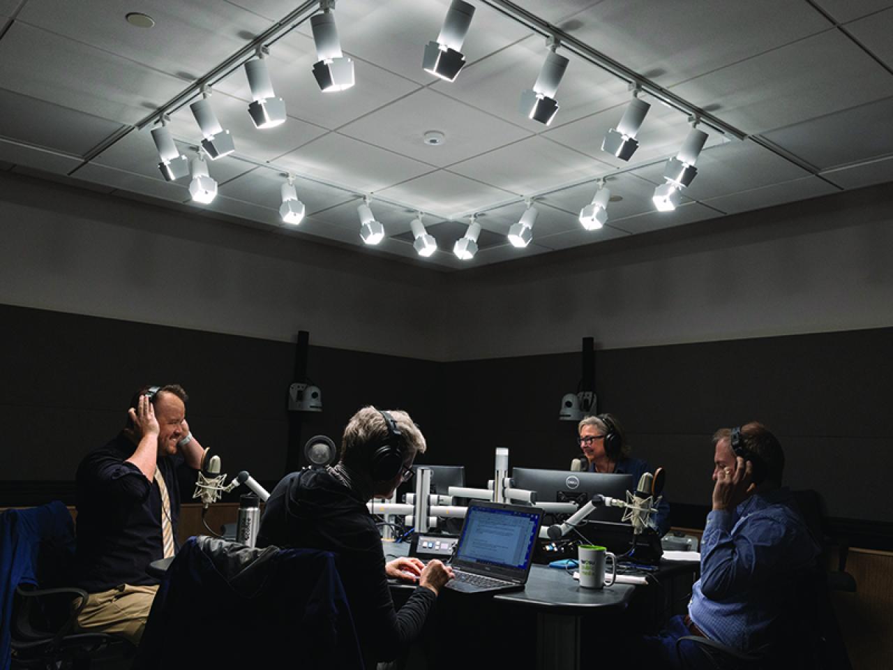 Radio studio with several people seated around a round table, wearing headphones and speaking into microphones under bright ceiling lights.