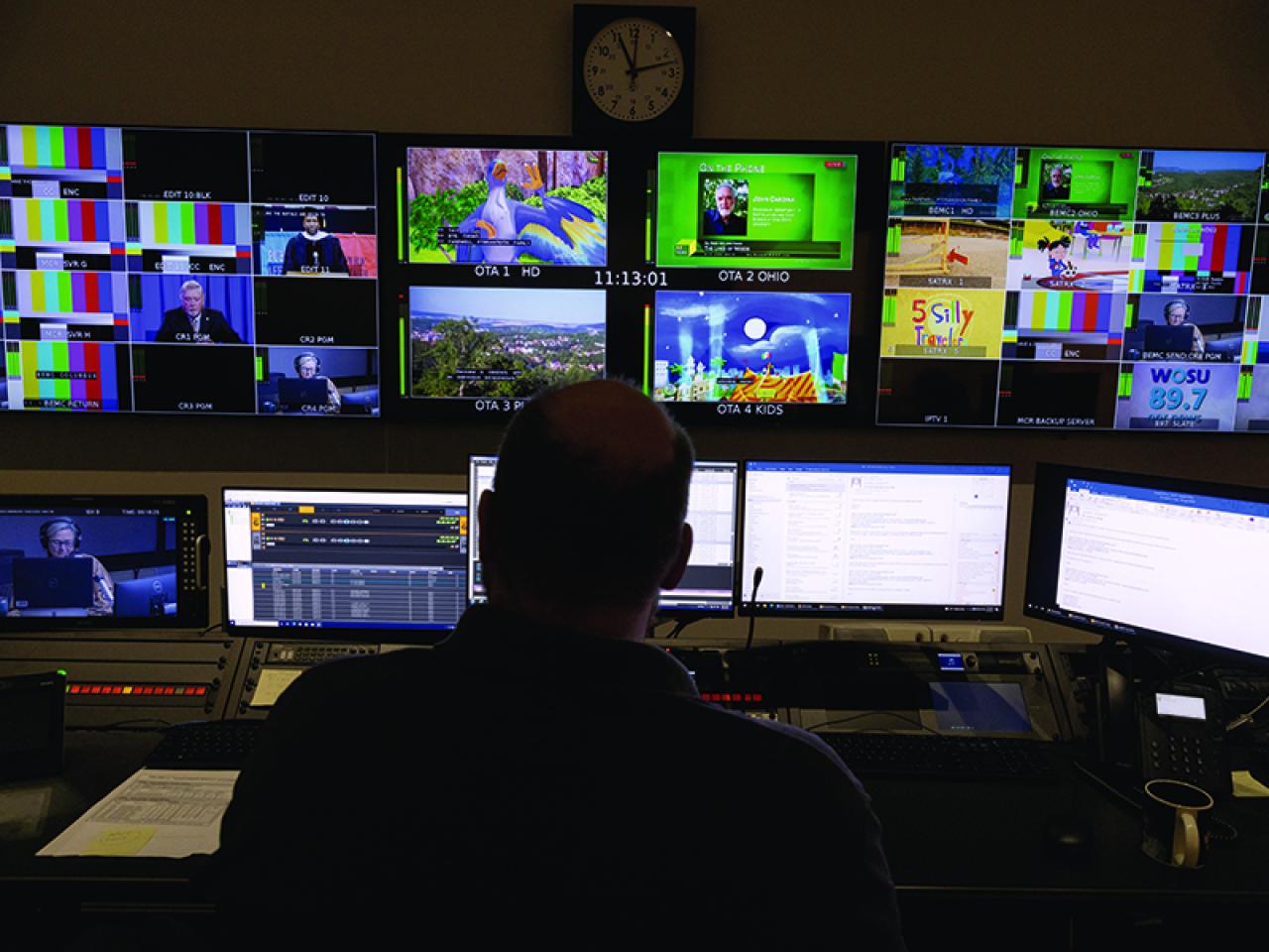 Broadcast control room with a person seated at a console facing multiple monitors showing live video feeds, color bars, and WOSU programming.