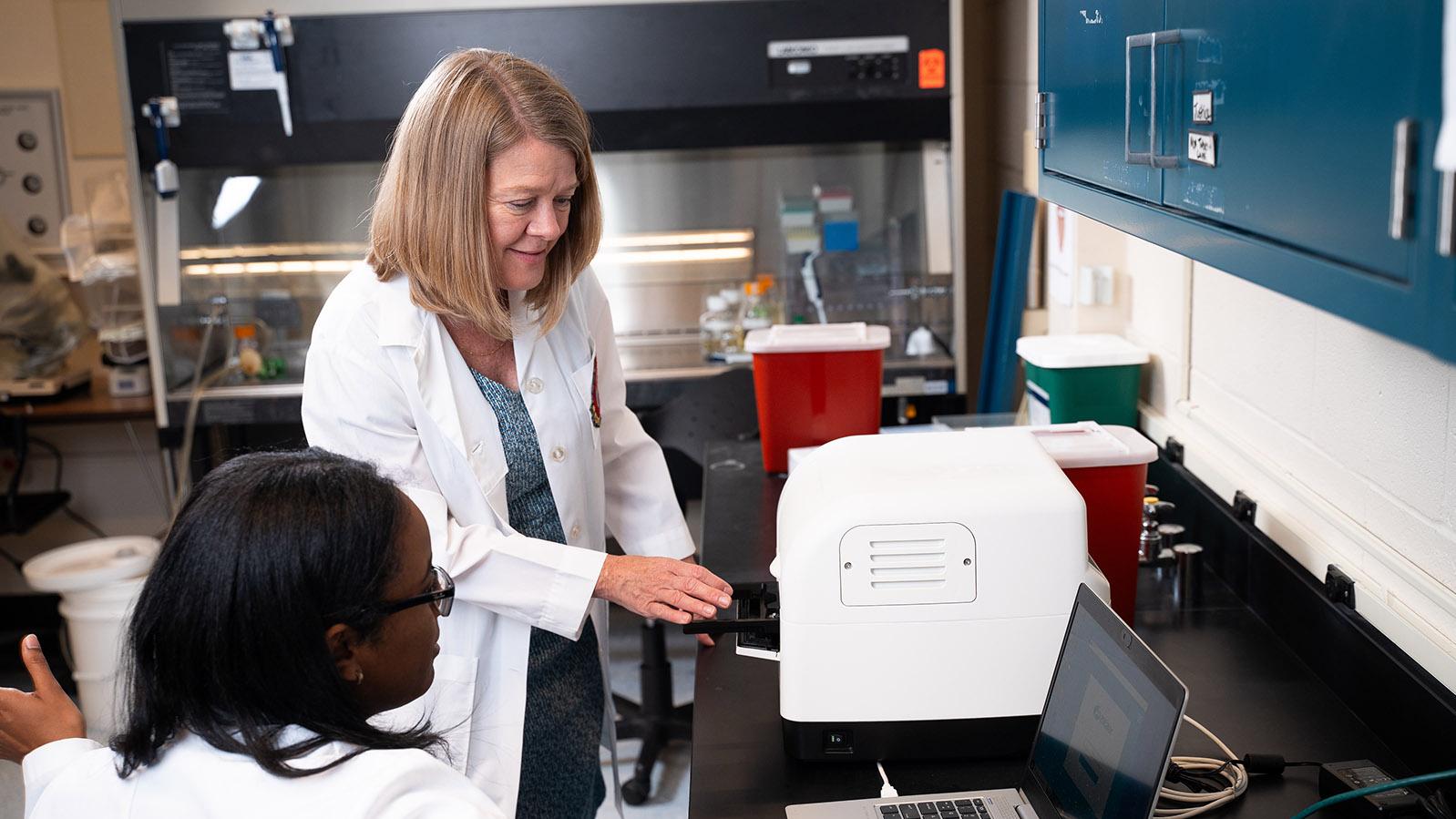 blonde-haired woman wearing a white lab coat speaks to a black-haired woman wearing a white lab coat