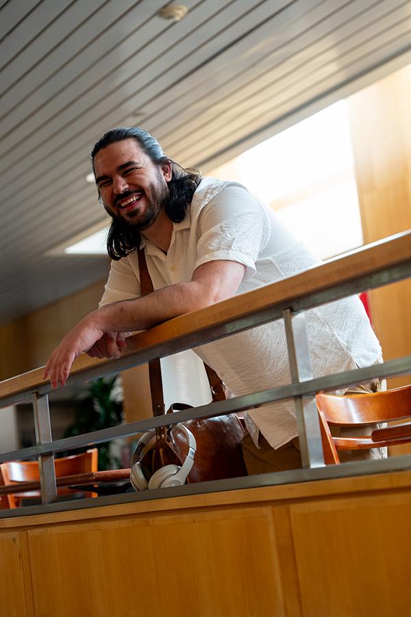 Levi leans on an indoor balcony railing, wearing a light short-sleeve shirt and carrying a brown shoulder bag