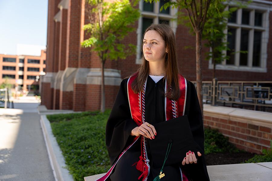 Olivia wearing a black cap and gown with red stole stands outdoors near a brick campus building