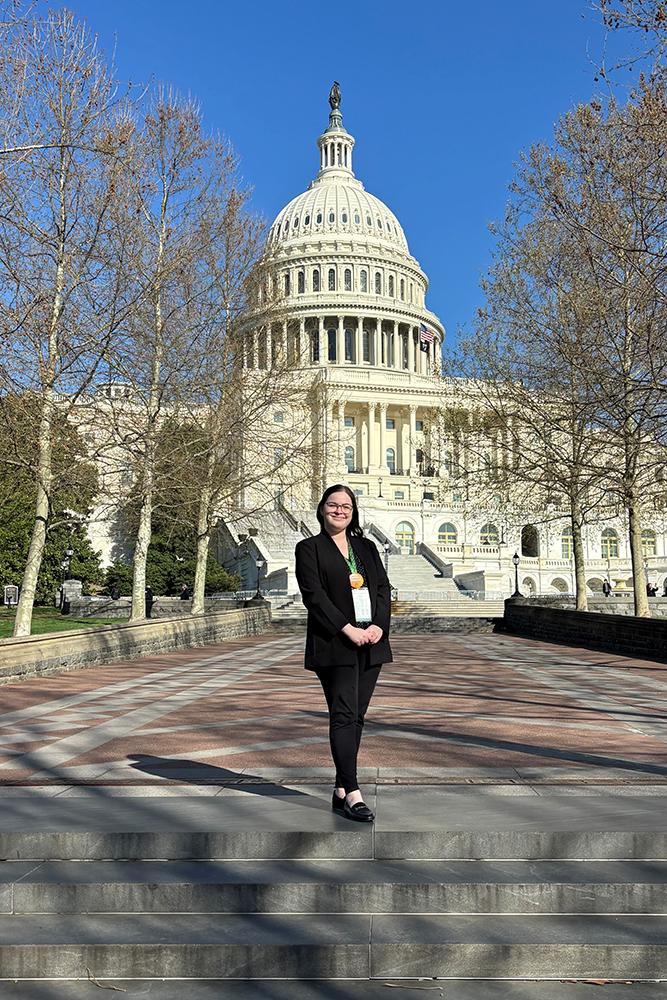 Ella stands on stone steps in front of the U.S. Capitol building, with the white dome centered behind them, leafless trees on both sides, and a clear blue sky overhead.