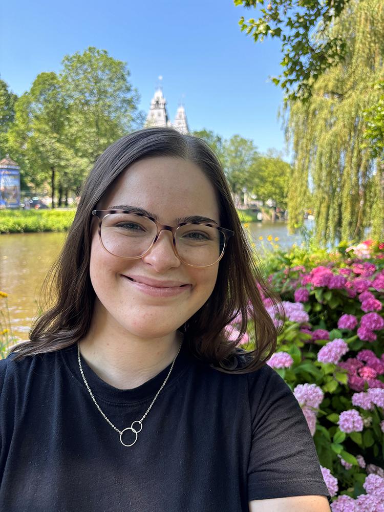 Ella takes a selfie by a canal in a park, with green trees, pink flowers, and historic buildings visible in the background under a clear blue sky.