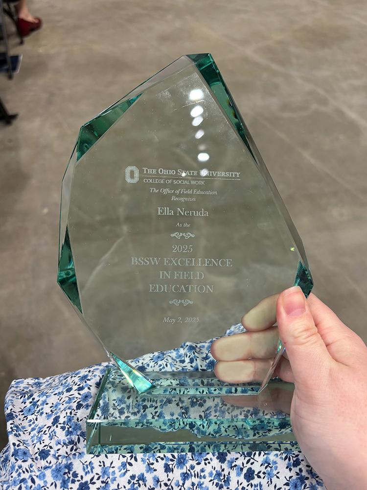 Ella's hand holds a clear glass award engraved with “The Ohio State University College of Social Work” and “2025 BSSW Excellence in Field Education,” resting on a floral-patterned dress in an indoor setting.
