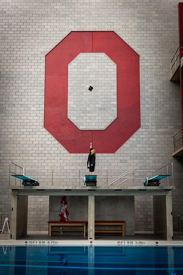 Lena stands on a diving platform, reaching upward, with a small object in the air, centered beneath a large red block “O” painted on a brick wall above an indoor swimming pool.