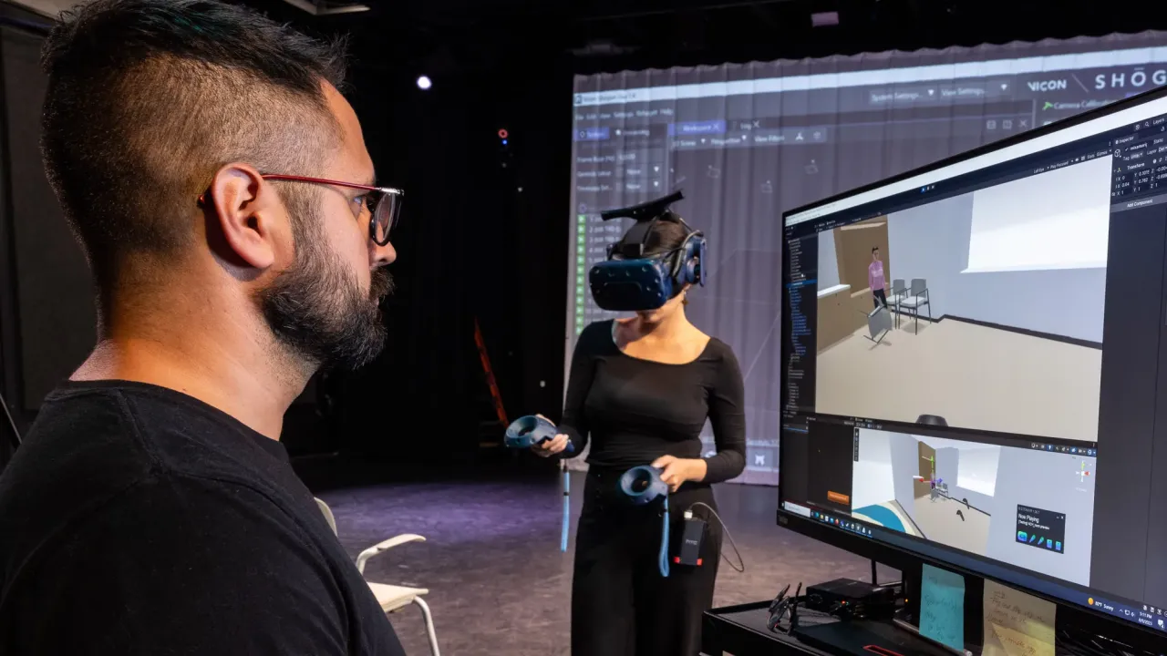 A man works on a computer whose screen shows a classroom setting while a woman on a stage beyond him wears a virtual reality headset and sensors used for motion capture.
