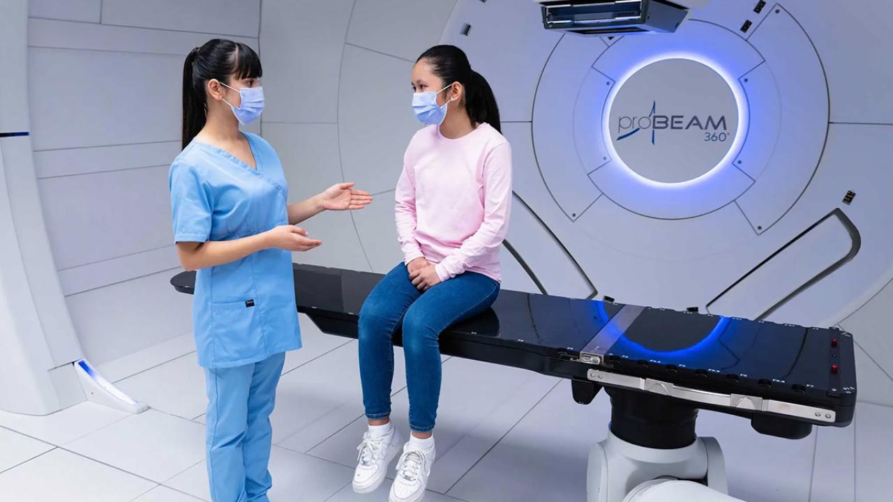 Medical staff member talking with a patient while they sit on the table of the FLASH Proton Therapy machine