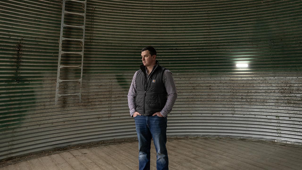 Will Hamman, a young white man wearing jeans and an insulated vest over his plaid button-down, stands inside an empty grain silo on his family farm in Ohio. The structure is corrugated metal maybe five times as wide as he is tall, with a peaked roof. A ladder and big flood light are too his right.