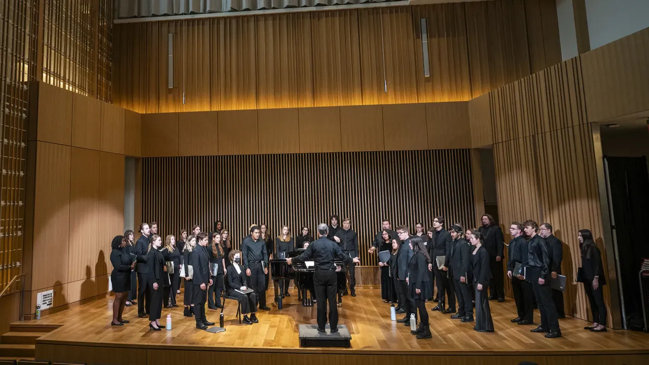 Students standing in rows wearing all black performance outfits singe on a wooden stage as a conductor holds his hands wide. The stage is wrapped in sound optimization and modern-looking elements. The lighting is very warm.