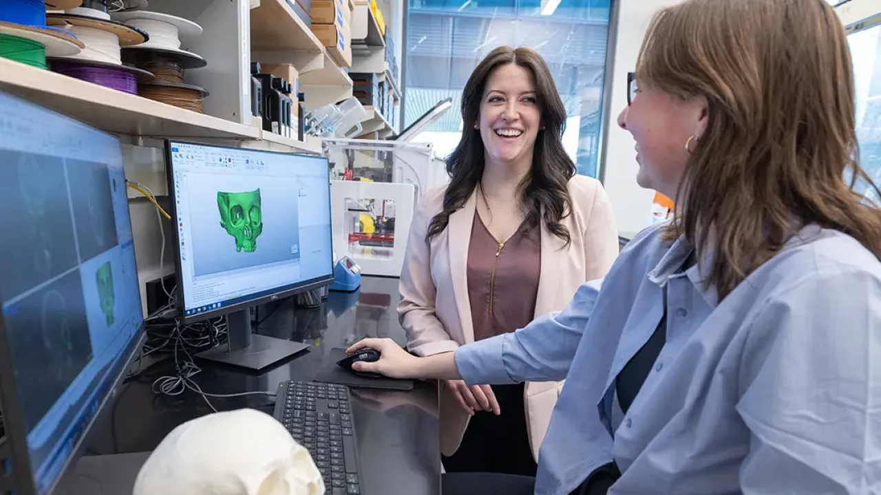 Two women talk in front on a computer monitor showing a 3-D skull. The 3-D printed result is on the table next to them.