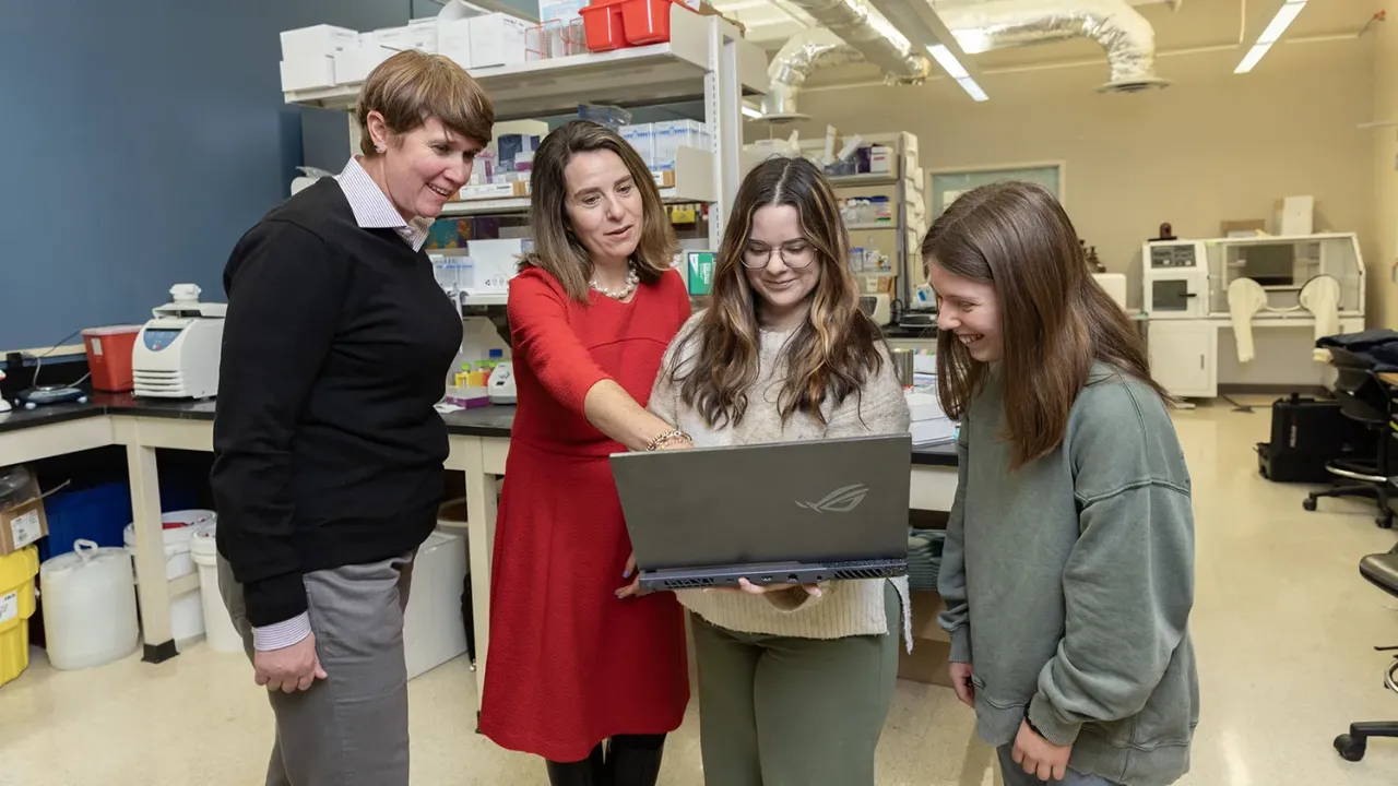 Dr. Tamar Gur, wearing a red dress, points at a laptop computer screen held by a young woman with long hair. Two more people are in the photo looking at the computer. They're all smiling and appear as though they enjoy working together.