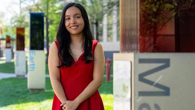 Hannah in a red sleeveless dress standing outdoors on a grassy campus area, with glass panels and signage in the background and trees around.